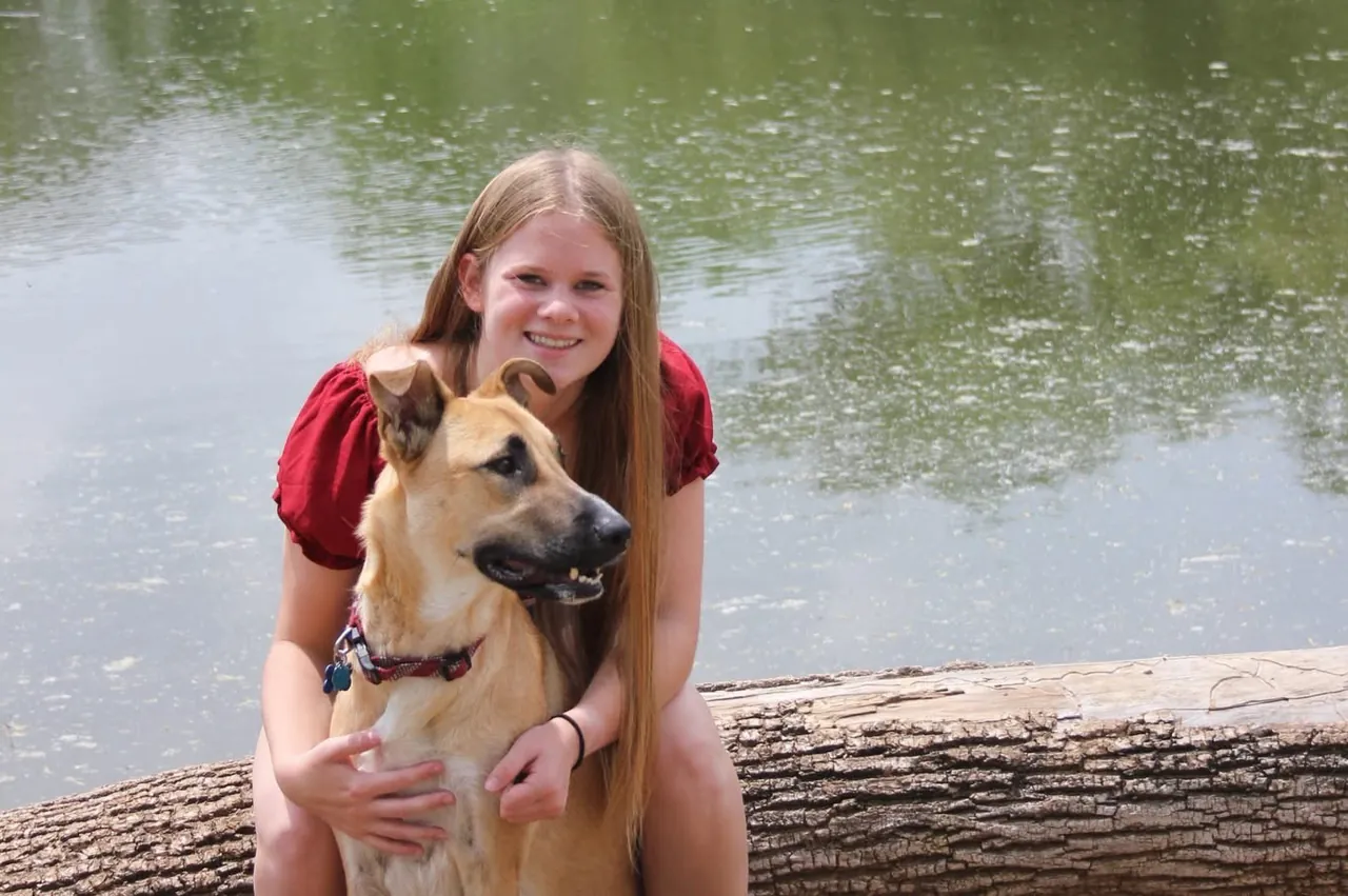Skylar Beun sitting on a log by a pond with her arms around a tan shepherd mix